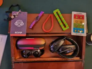 A lanyard, fidget toys, stickers with moods, colorblind glasses, and ear defenders on a wooden box on a green tablecloth