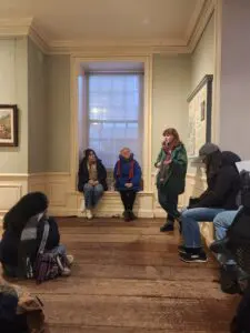 Woman standing, speaking to a group of people sitting on a windowsill, with one person sitting on the floor