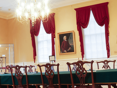 A historic room with red curtains, a chandelier, and a long table with chairs. A portrait hangs on the wall.