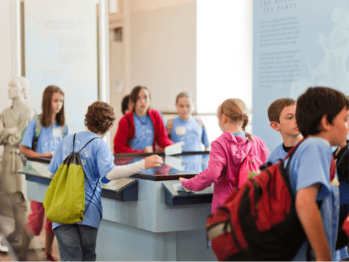 A group of children in a museum setting, possibly on a school field trip, exploring exhibits with a guide.