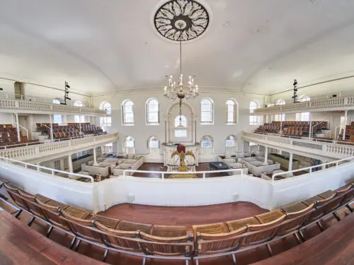 Panoramic view of the interior of Boston's Old South Meeting House, showing the historic meeting hall with its central pulpit, wraparound galleries, chandeliers, and wooden pews.