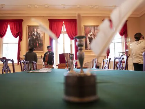 A historical room with red curtains, portraits, a green table, and visitors.