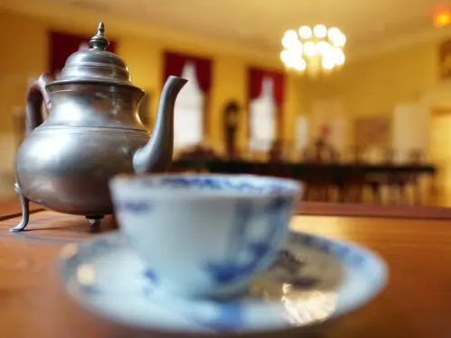 Close-up of a teacup, saucer, and metal teapot, with a long table, tall windows, and chandelier out of focus in the background