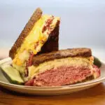 A Reuben sandwich with corned beef, cheese, and a pickle slice on the side.