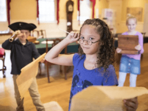 Children in a historical setting engaged in a playful activity, wearing costumes and holding maps or papers.