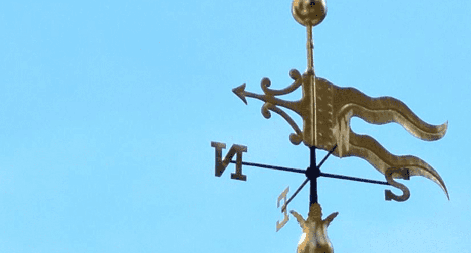 Golden weathervane with ornate flag design against a light blue background