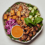 A bowl with tofu, mushrooms, cucumbers, red cabbage, greens, and a side of sauce.