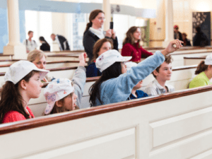Students are seated in a historical setting, some raising hands, engaged in a Boston Tea Party experience.