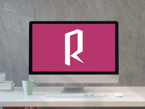 A computer monitor displaying a stylized "R" on a pink background, sitting on a desk with a keyboard, mouse, and a green mug.