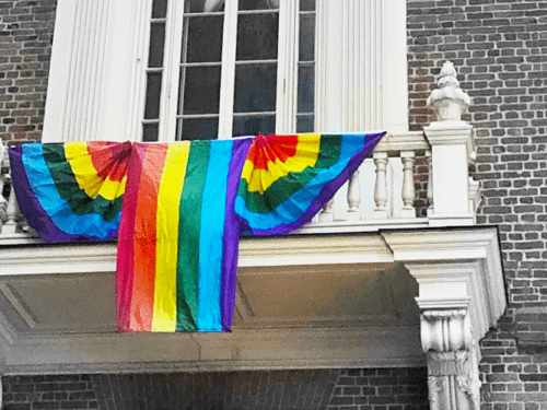 Black-and-white photo of an ornate balcony on a brick building, draped with banners and a pride flag that have been edited to appear in full color