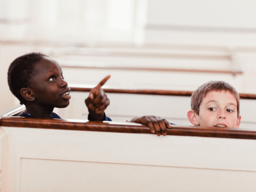 Two children are inside a chapel, with one pointing and the other looking over a pew.