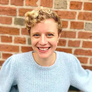 A person with short curly hair, wearing a light blue sweater, smiles in front of a brick wall.