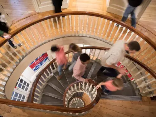 A spiral staircase with people moving up and down. Sign reads "Explore". This is related to accessibility at revolutionary spaces.
