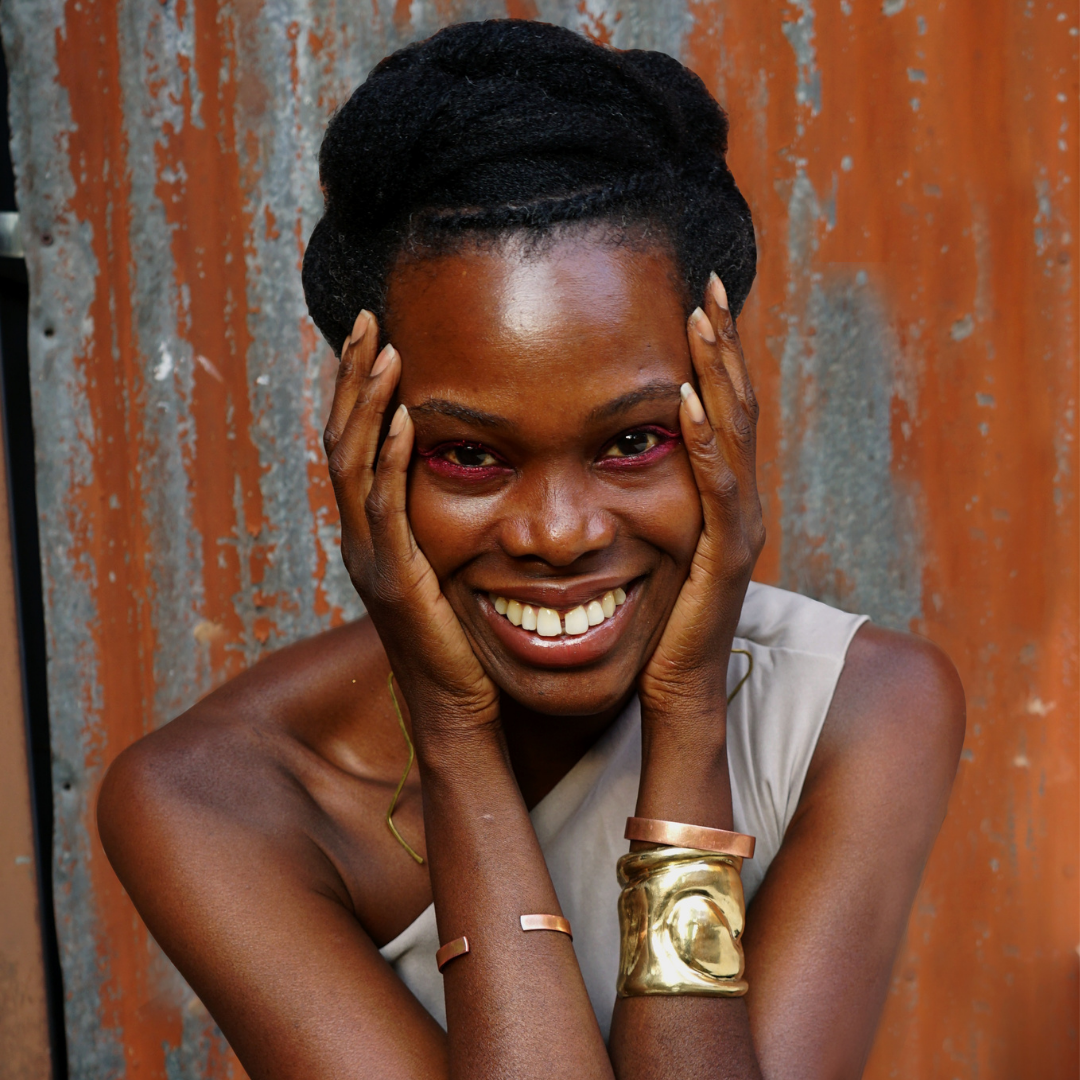 A woman with an elegant hairstyle and gold jewelry smiles against a textured, rustic backdrop.