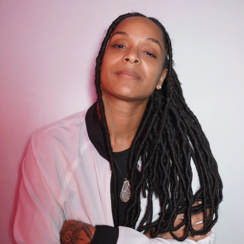 A person with long hair and a pendant, wearing a white jacket over a black top, stands confidently with arms crossed.