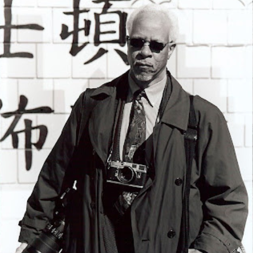 A man wearing sunglasses and a coat with a camera around his neck stands in front of a wall with large Asian characters.